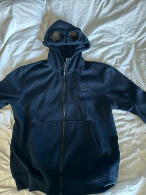 Svart hoodie med goggles från C.P. Company - Säljer en svart hoodie från C.P. Company med dragkedja och ikoniska goggles i huvan. Hoodien har två stora fickor framtill och broderad logga på bröstet. Perfekt för dig som vill sticka ut med en unik streetwear-look. Använd fåtal gånger, nytt skick 