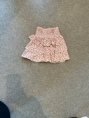 Rosa blommig volangkjol - Supersöt kort kjol med rosa blommigt mönster och volanger. Kjolen har smock i midjan och är perfekt för varma dagar. Lätt och luftig känsla, passar till både sneakers och sandaler.