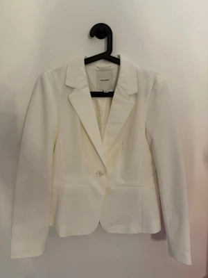 Vit kavaj från Vero Moda - Stilren vit kavaj från Vero Moda med klassisk krage och en knapp framtill. Kavajen har två fickor och är figursydd för en snygg siluett. Perfekt att styla med jeans eller kjol för en trendig look.