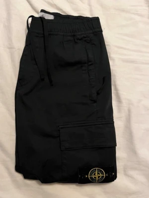 Svarta cargobyxor från Stone Island - Säljer ett par svarta cargobyxor från Stone Island med klassisk patch på benet. Byxorna har flera fickor, resår i midjan med dragsko och muddar vid bensluten. Uppsydda för att passa 169-174 cm perfekt. Sparsamt använda, givetvis äkta. CLG-kod finns. 