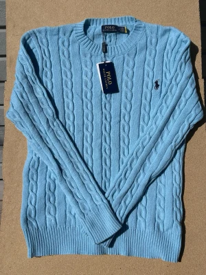 Ljusblå kabelstickad tröja Polo Ralph Lauren - Ljusblå kabelstickad tröja från Polo Ralph Lauren med rund hals och broderad logotyp på bröstet. Tröjan har ribbade muddar vid ärmslut och nederkant, och är tillverkad i mjukt material. Perfekt för dig som gillar klassisk och stilren design.