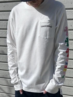 Vit långärmad topp från Chrome Hearts - Vit långärmad topp från Chrome Hearts med färgglada blommönster på ryggen och ärmarna. Bröstficka med logotyp och tryck på ryggen. T-shirten har en relaxed passform och är tillverkad i mjuk bomull.