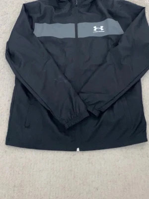Svart vindjacka från Under Armour - Svart vindjacka från Under Armour med grått band och logga över bröstet. Jackan har dragkedja framtill, elastiska muddar och två sidofickor. Perfekt för blåsiga dagar och sportiga outfits.