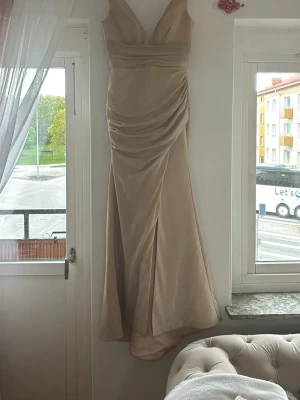 Beige långklänning med snörning - Elegant beige långklänning med v-ringning och draperad framsida. Klänningen har en slits framtill och snörning i ryggen för en justerbar passform. Tillverkad i ett mjukt, glansigt material som ger en lyxig känsla. Perfekt för speciella tillfällen.