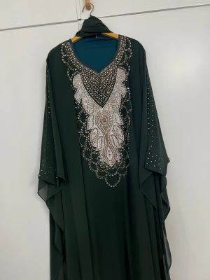 Mörkgrön kaftanklänning med pärlor - Elegant mörkgrön kaftanklänning med v-ringning och detaljerad pärl- och paljettbrodyr framtill. Vida ärmar med små guldfärgade nitar och matchande sjal. Flödande passform i lätt, syntetiskt material.
