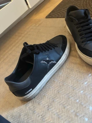 Svarta Axel Arigato sneakers EU 44 nypris: 2750kr - Svarta sneakers från Axel Arigato med vit sula och svart snörning. Skorna har en fågelbrodyr på sidan och texten '140MM' vid hälen. Tillverkade i läder med detaljer i mocka på tån och hälen. Snygg och stilren design som passar till många outfits.