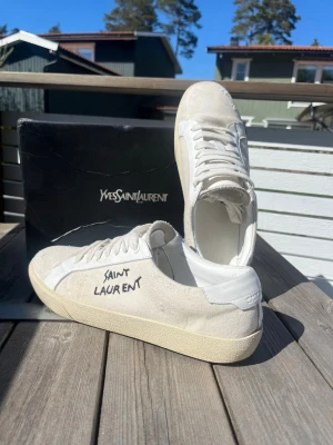 Saint Laurent vita sneakers - Snygga vita sneakers från saint laurent. Skorna har en klassisk låg profil, ovandel i mocka och läder samt vita snören och sula. Perfekta för dig som gillar stilrena och exklusiva sneakers med en unik touch. Perfekta skor till sommaren, skriv vid minsta fundering. Köpte dem från Plick förra sommaren säljer pågrund av att dem är för små.