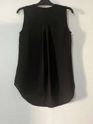 Svart ärmlös blus från Broadway - Stilren svart ärmlös blus från Broadway NYC Fashion. Blusen har en lös passform, lätt v-ringning och knäppning i nacken. Perfekt att matcha med jeans eller kjol för en snygg look. Obs saknar knappen