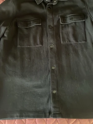 Svart overshirt med tryckknappar - Stilren svart overshirt med två stora bröstfickor med lock och tryckknappar framtill. Klassisk krage och rak passform. Perfekt att slänga över en hoodie eller t-shirt för en cool look.