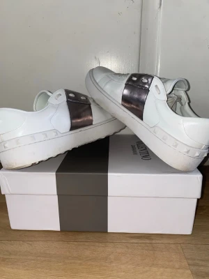 Valentino Garavani sneakers med metallic - Snygga vita sneakers från Valentino Garavani med en bred metallic-silver rem över sidan och coola nitar på hälen. Skorna har snörning och en platt sula, tillverkade i skinn för en lyxig känsla. Perfekt för dig som gillar statement-detaljer och exklusiv design.