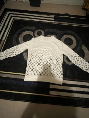 Vit Louis Vuitton sweatshirt - Snygg vit sweatshirt från Louis Vuitton med klassiskt LV-monogram i svart över hela tröjan. Tröjan har rund halsringning, långa ärmar och ribbade muddar. Perfekt för dig som gillar exklusiv streetwear och vill sticka ut med en ikonisk design.