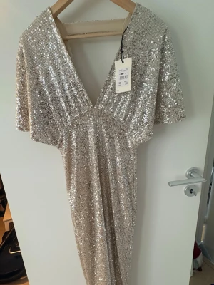 Glittrig långklänning i champagne - Snygg långklänning i champagnefärg med paljetter över hela klänningen. Djup v-ringning och korta ärmar ger en trendig look. Perfekt för dig som vill sticka ut med en glittrig och elegant stil.