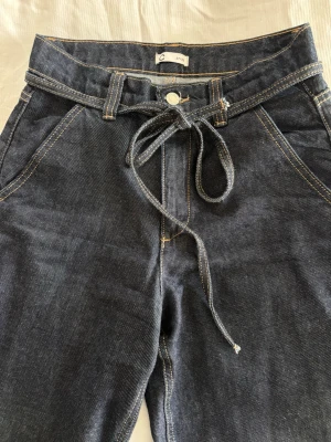 Mörkblå jeans med knytskärp - Snygga mörkblå jeans från Cubus med raka ben och knytskärp i midjan. Jeansen har kontrastsömmar och djupa fickor framtill. Perfekta för en avslappnad och trendig look. Materialet är klassisk denim i bomull.