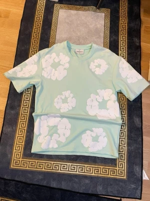 Mintgrön t-shirt med vita blommor - Fräsch mintgrön t-shirt från Hul le Kes med stora vita blommiga mönster över hela plagget. T-shirten har rund hals, korta ärmar och är gjord i mjuk bomull. Perfekt för dig som vill sticka ut med en unik och färgglad stil.