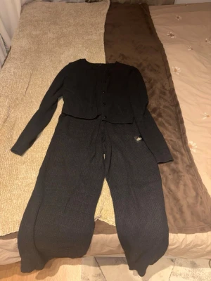 Svart ribbat loungewear-set från Alo - Mysigt svart loungewear-set från Alo med ribbad struktur. Setet består av en långärmad kofta med knappar och matchande vida byxor med dragsko i midjan. Båda delarna har Alo-logga framtill. Perfekt för chill dagar hemma.