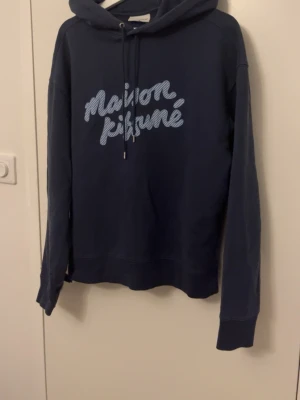 Blå Maison Kitsuné hoodie - Mörkblå hoodie från Maison Kitsuné med stor vit logga i skrivstil på bröstet. Klassisk huva med dragsko och långa ärmar. Tillverkad i mjukt bomullsmaterial som känns skönt mot huden. Perfekt för en avslappnad och trendig look.