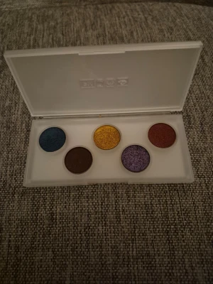 Färgstark ögonskuggspalett PM - Snygg ögonskuggspalett från Pat McGrath Labs med fem intensiva färger: blå, guld, röd, brun och lila. Paletten har en stilren, matt vit plastförpackning med genomskinligt lock och guldiga detaljer. Perfekt för dig som gillar att experimentera med färg. Ny inte använd. 