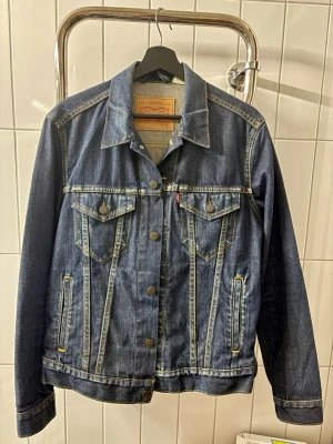 Levi's klassisk blå jeansjacka S - Säljer en klassisk blå jeansjacka från Levi's med snygga kontrastsömmar och två bröstfickor med knappar. Jackan har en tidlös design, knappstängning framtill och ikonisk Levi's-lapp i nacken. Perfekt för dig som vill ha en stilren och ikonisk look. Aldrig använt!                 Nypris 1499kr