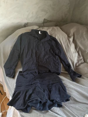 Svart skjorta med volangkjol - Ett linne det. Säljer en svart skjorta med klassisk krage och knäppning framtill. Till skjortan hör en matchande svart kjol med volangdetaljer och veckade partier upptill. Setet har en avslappnad passform och är tillverkat i ett mjukt tyg, perfekt för dig som gillar stilrena och enkla plagg. Storlek 38, från Nelly 