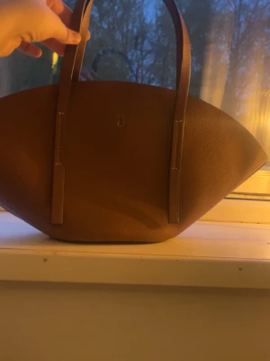 Brun handväska i läderlook - Snygg brun handväska med två långa handtag och stilren, minimalistisk design. Väskan har en unik, bred form och är tillverkad i ett material som liknar läder. Perfekt för dig som gillar trendiga accessoarer med enkel look.