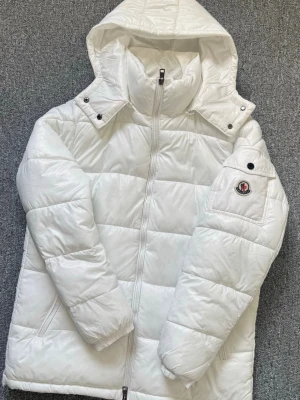 Vit dunjacka från Moncler  - Moncler Maya jacka i vit, NFC märke tillgängligt. Jackan har Moncler-logga på ärmen, två sidofickor och quiltad design. 