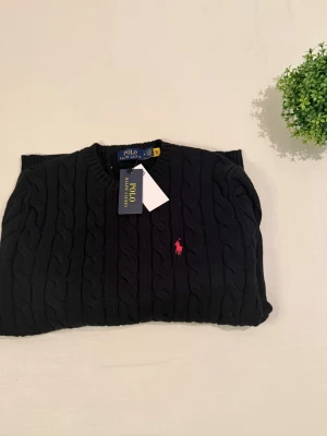 Svart kabelstickad tröja Polo Ralph Lauren - Kabelstickad svart tröja från Polo Ralph Lauren med rund halsringning och röd broderad logga på bröstet. Tröjan har långa ärmar och ribbade muddar vid ärmslut och nederkant. Perfekt för en stilren och klassisk look.