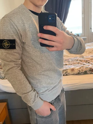 Grå sweatshirt  - Grå sweatshirt från Stone Island med klassisk rund hals och ribbade muddar. Ikonisk svart logotyp-patch på vänster ärm. Perfekt för en stilren och avslappnad look. Materialet är mjukt och bekvämt bomullstyg.