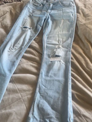Ljusblå ripped jeans med raka ben - Säljer ett par ljusblå jeans med raka ben och slitna detaljer på båda benen. Jeansen har klassisk femficksdesign och är tillverkade i denim. Perfekta för en avslappnad och trendig look.