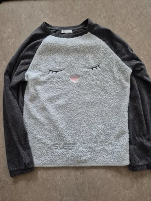 Tröja - Mysig långärmad tröja i grått med fluffig, ljus framsida och ett broderat sömnigt ansikte samt liten rosa nos. Ärmar och baksida är i mjuk, mörkgrå fleece. Perfekt för chill dagar.