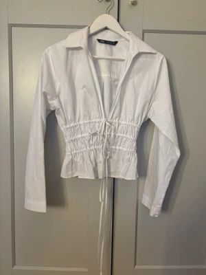 Vit blus med dragsko från Zara - Snygg vit blus från Zara med krage och långa ärmar. Blusen har markerad midja med dragsko och volangkant nedtill. Perfekt för en trendig och stilren look. Tillverkad i ett lätt bomullstyg.