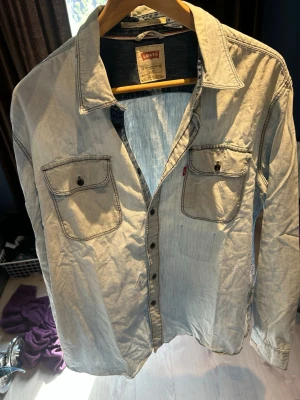 Ljusblå jeansskjorta från Levi's - Säljer en klassisk ljusblå jeansskjorta från Levi's med två bröstfickor och svarta knappar. Skjortan har lång ärm och en avslappnad passform, perfekt för en cool och enkel stil. Ikonisk röd Levi's-tagg på ena fickan.