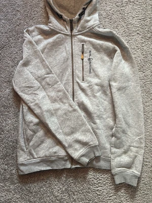 Grå hoodie från Sail Racing - Snygg grå hoodie från Sail Racing med dragkedja framtill och broderad logga på bröstet. Har en praktisk bröstficka med dragkedja och justerbar huva med snören. Tillverkad i mjukt material och har ribbade muddar vid ärmslut och nederkant.