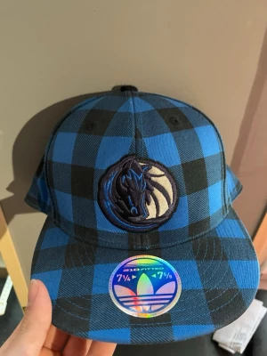 Blårutig keps Adidas x Mavericks - Säljer en riktigt snygg keps från Adidas med blå och svart rutig design och Dallas Mavericks-logga broderad framtill. Kepsen har Adidas Originals-logga baktill och är en fitted modell. Perfekt för dig som gillar sportig streetstyle och vill sticka ut.