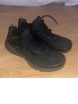 Svarta Nike Presto sneakers - Svarta Nike Presto sneakers med låg profil och klassisk siluett. Skorna har en ovandel i mesh och syntet, med svarta skosnören och diskret Nike-logga på sidan och tungan. Perfekta för en clean och sportig look sko storlek 44.