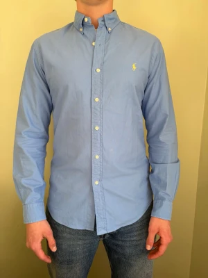 Blå skjorta från Ralph Lauren - Klassisk ljusblå skjorta från Ralph Lauren med button-down krage och broderad logga på bröstet. Skjortan har långa ärmar, normal passform och är tillverkad i bomull. Perfekt till jeans eller chinos.