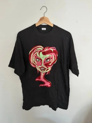 Svart t-shirt cool vintage street  - Cool svart t-shirt med en stor, färgstark grafisk illustration av ett ansikte i rött, rosa och grönt på framsidan. Storlek L