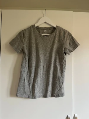 Grå basic t-shirt från 157 - Enkel och stilren grå t-shirt från 157 i storlek XS. T-shirten har rund halsringning och korta ärmar. Tillverkad i mjuk bomullsblandning med 90% bomull och 10% viskos. Perfekt till jeans eller shorts för en avslappnad look.