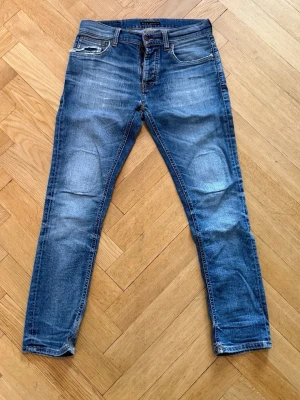 Blå jeans från Nudie Jeans - Snygga blå jeans från Nudie Jeans med slitningar och en trasig detalj vid myntfickan. Storlek 30/30. Modell Grim Tim. Klassisk femficksmodell med dragkedja och knapp. Jeansen har en tvättad look och smal passform, perfekta till sneakers eller boots. 