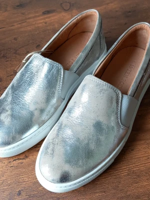 Silvriga slip-on sneakers från Ten Points storlek 39 - Slip-on sneakers i metallic-silver från Ten Points. Skorna har en låg profil, elastiska paneler på sidorna och vit sula. Ovandelen är i glansigt läder material och insidan är fodrad i ljusbrunt läder. Perfekt för dig som gillar en trendig och stilren look.
