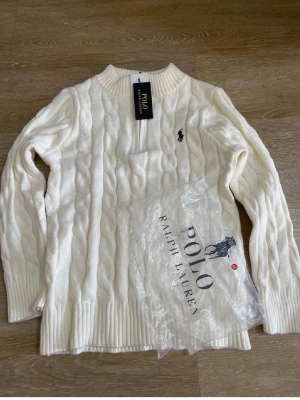 Vit kabelstickad tröja Polo Ralph Lauren - Vit kabelstickad tröja från Polo Ralph Lauren med rund halsringning och svart broderad logga på bröstet. Tröjan har långa ärmar och ribbstickade muddar. Klassisk och stilren design i mjukt material.