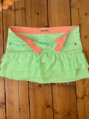 Lågmidjad volangkjol neon - Supercool neongrön kjol, märke Terranova, med  tre volanger. Kjolen har fickor fram och en snygg kontrastfärgad insida i orange. Perfekt för dig som vill sticka ut med en färgglad stil. Noggrant omsydda jeans. 