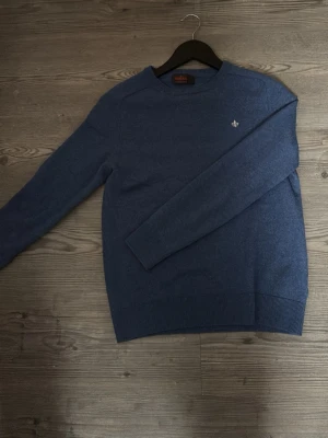 Blå stickad tröja Morris - Snygg blå stickad tröja från Morris med rund halsringning och diskret broderad logga på bröstet. Tröjan har långa ärmar och ribbade muddar vid ärmslut och nederkant. Perfekt för lager på lager och enkel att matcha.