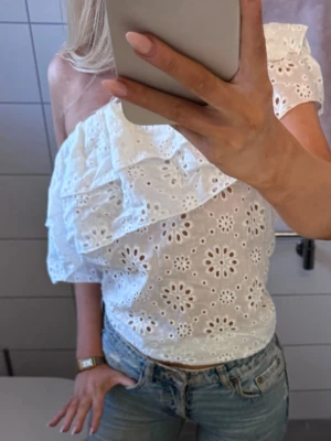 Vit oneshoulder blus med broderi - Superfin vit blus med oneshoulder och volangdetalj. Blusen har broderade blommor och är perfekt för dig som gillar romantisk stil. Luftigt material som passar bra till både jeans och kjol.
