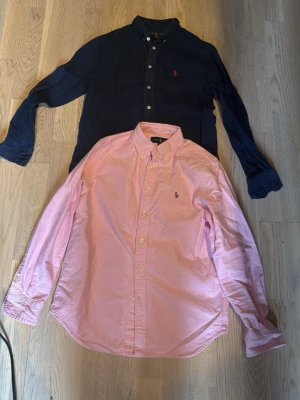 Ralph Lauren skjortor i rosa och marinblå - Två klassiska skjortor från Ralph Lauren, en i ljusrosa och en i marinblå. Båda har lång ärm, knappar framtill och broderad logga på bröstet. Perfekta för en stilren och tidlös look. 250 styck men om du köper båda får du paketpris på 400!! Storlek 10-12år i barn!!