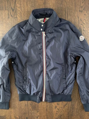 Moncler Windbreaker Bomberjacket - Selling a Moncler windbreaker in very good condition. It hasn’t been worn for a while which makes it very well preserved.  Lightweight and practical jacket with a clean design, perfect for spring. High quality as expected from Moncler. No receipt but it is authentic. Condition: Very good Usage: Lightly worn Length : 57 cm Size is 13-15 years old  Original price : around 7000 SEK Price : 3000 SEK