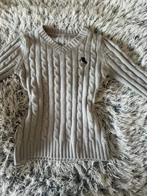 Grå kabelstickad tröja Polo Ralph Lauren - Aldrig använt. Snygg grå kabelstickad tröja från Polo Ralph Lauren med V-ringning och svart broderad logga på bröstet. Tröjan har långa ärmar och ribbade muddar vid ärmslut och nederkant. Perfekt för lager-på-lager och klassisk stil.