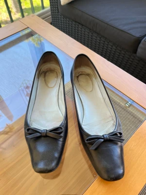 Svarta ballerinaskor från Ross. Storlek 37 - Svarta ballerinaskor med rosett framtill. Skorna har en platt sula och en rundad tå. Insidan är ljus och sulan är svart. Skinn (nappa). Storlek 37. 