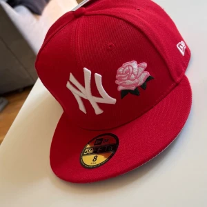Röd New Era Yankees keps med ros - Snygg röd keps från New Era med New York Yankees-logga och broderad ros framtill. Kepsen har platt skärm, broderad 1999 World Series-patch på sidan och klassisk 59FIFTY-stil. Perfekt för dig som vill sticka ut med en unik accessoar.