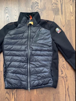  Parajumper cardigan  - Snygg svart pufferjacka från Parajumpers med quiltad front och rygg, samt släta svarta ärmar. Jackan har dragkedja framtill, bröstficka med dragkedja och klassisk Parajumpers-logga på ärmen. Perfekt för kalla vinterdagar.