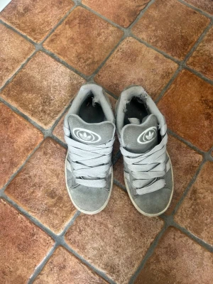 Gråa Adidas high top sneakers - Gråa high top sneakers från Adidas med breda skosnören och klassisk Adidas-logga på plösen. Skorna är i mockaliknande material och har en vit sula som kontrast. Perfekta för en streetwear-look.